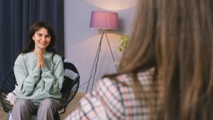Positive young woman in casual clothes smiling while sitting on chair after psychotherapy session with anonymous female doctor in modern office